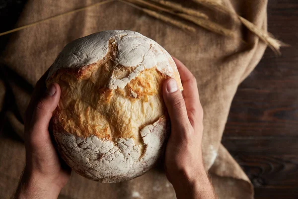 Cropped shot of male baker holding bread over wooden table with wheat and sackcloth — Stock Photo