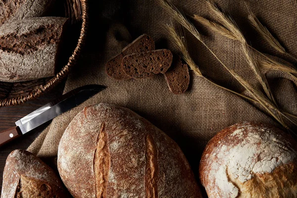 Elevated view of various types of bread, knife, wicker breadbasket on sackcloth — Stock Photo