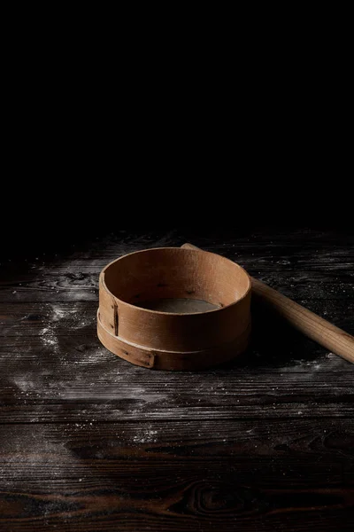 Vista de cerca del tamiz y el rodillo en la cubierta de mesa de madera por harina aislada sobre fondo negro - foto de stock