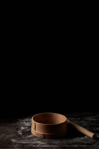 Closeup shot of sieve and rolling pin on wooden table covering by flour isolated on black background — Stock Photo