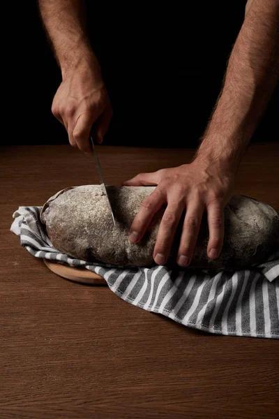 Vue partielle du boulanger mâle coupant le pain au couteau sur un sac sur une table en bois — Photo de stock