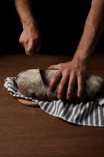 Partial view of male baker cutting bread by knife on sackcloth on wooden table — Stock Photo