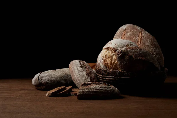 Closeup shot of various types of bread and wicker breadbasket on wooden table isolated on black background — Stock Photo