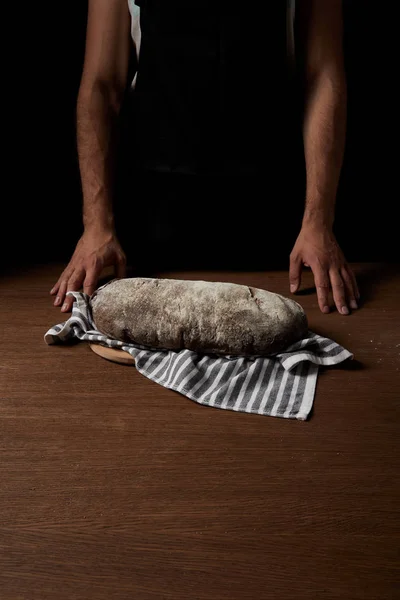 Vue partielle du boulanger mâle dans le tablier debout près de la table avec planche à découper, sac et pain — Photo de stock