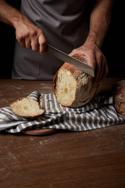 Vista parziale del panettiere maschio che taglia il pane con un coltello sul sacco sul tavolo di legno — Foto stock
