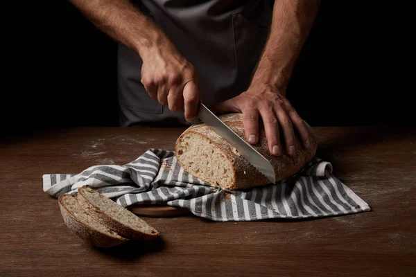 Cropped shot of male baker cutting bread by knife on sackcloth on wooden table — Stock Photo