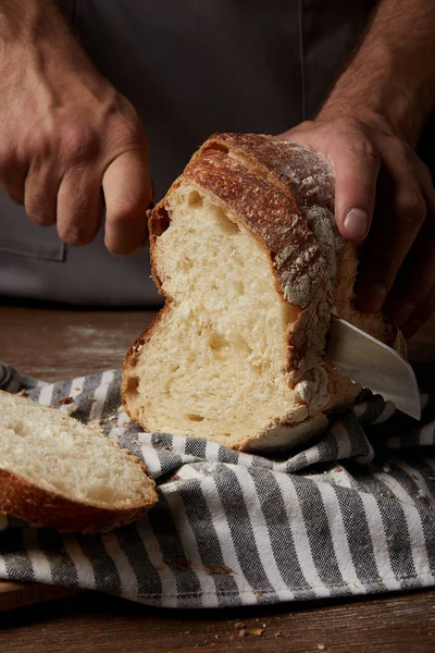 Cropped shot of male baker cutting bread by knife on sackcloth on wooden table — Stock Photo