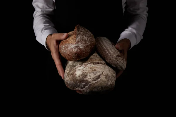 Visão parcial do padeiro macho em avental segurando vários tipos de pão isolado no fundo preto — Fotografia de Stock