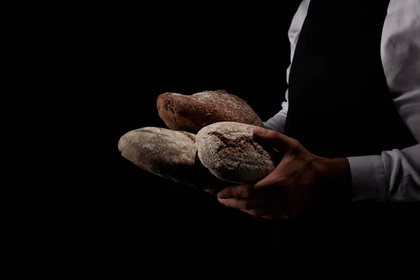 Immagine ritagliata di panettiere maschio in grembiule contenente vari tipi di pane isolato su sfondo nero — Foto stock