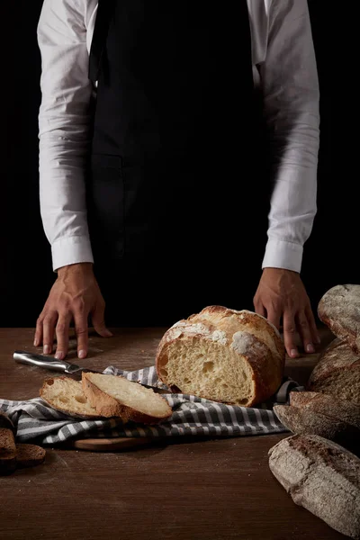 Imagen recortada de panadero masculino en delantal de pie cerca de la mesa con pan, cuchillo y saco - foto de stock