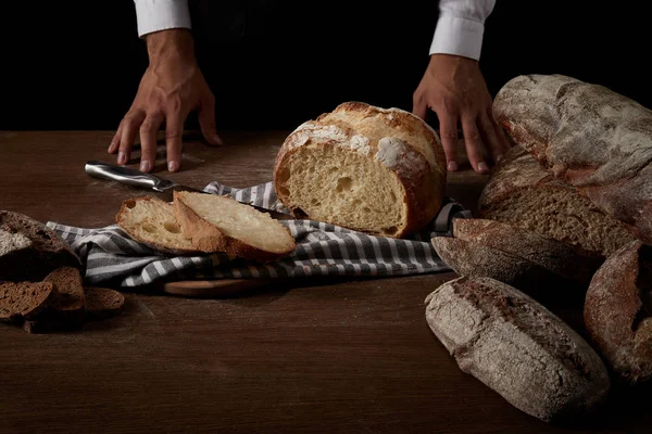 Частичный вид пекаря-мужчины, стоящего рядом со столом с хлебом, ножом и мешковиной — стоковое фото