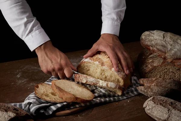 Vista parcial del panadero macho cortando pan por cuchillo sobre tela de saco sobre mesa de madera - foto de stock