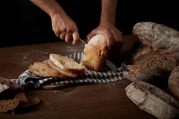 Cropped shot of male baker cutting bread by knife on sackcloth on wooden table — Stock Photo