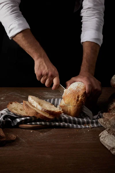 Partial view of male baker in apron cutting bread by knife on sackcloth on wooden table — Stock Photo