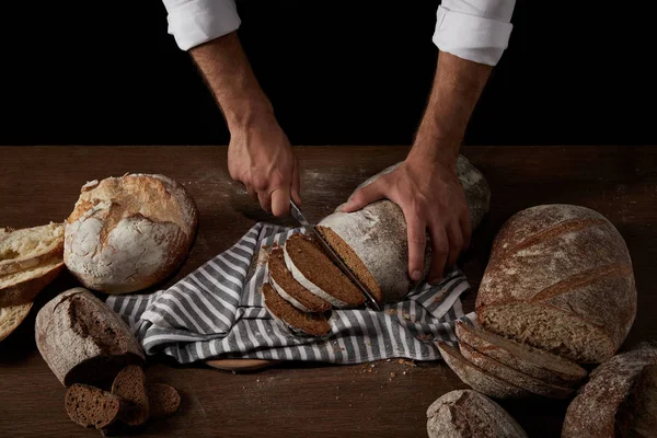 Immagine ritagliata del panettiere maschio che taglia il pane con un coltello sul sacco sul tavolo di legno — Foto stock