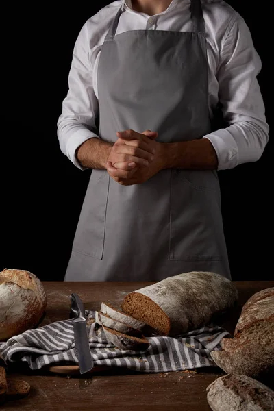 Tiro recortado de panadero masculino en delantal de pie cerca de la mesa con cuchillo, saco y pan - foto de stock