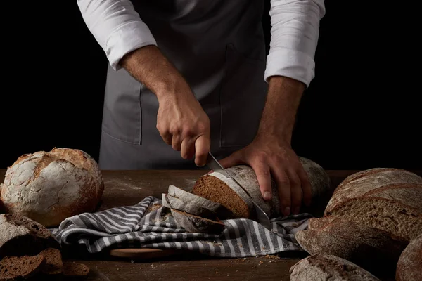 Vista parziale del panettiere maschio in grembiule che taglia il pane sul sacco con un coltello sul tavolo di legno — Foto stock