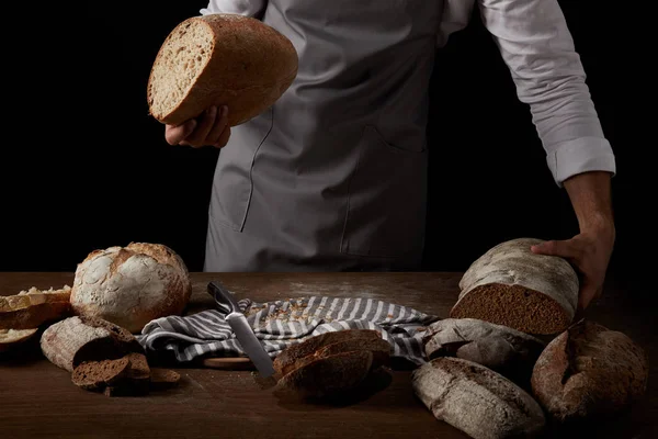 Partial view of male baker in apron holding bread over table with knife, bread and sackcloth — Stock Photo