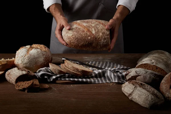 Cropped image of male baker in apron holding bread over table with knife, bread and sackcloth — Stock Photo