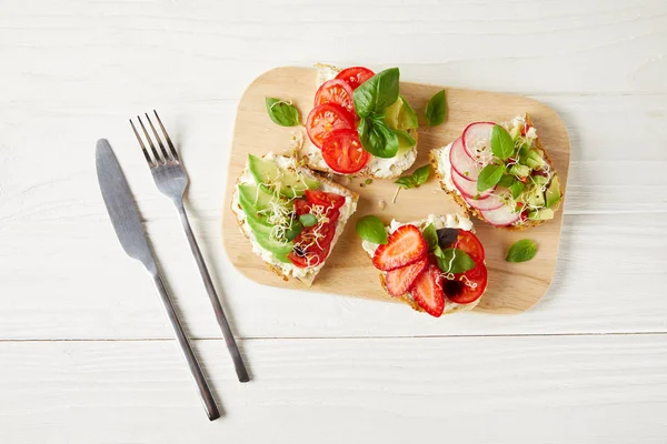 Top view of various sandwiches on cutting board with cutlery on white wooden surface — Stock Photo