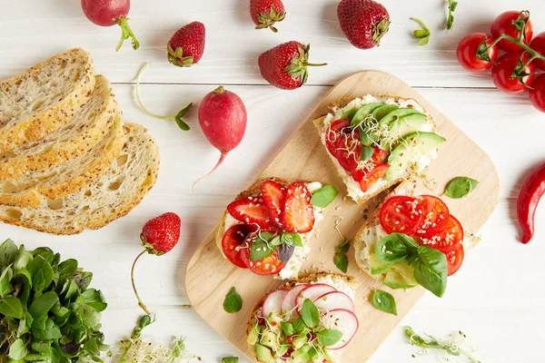 Vista superior de sabrosos sándwiches en la tabla de cortar rodeada de ingredientes en la superficie de madera blanca - foto de stock