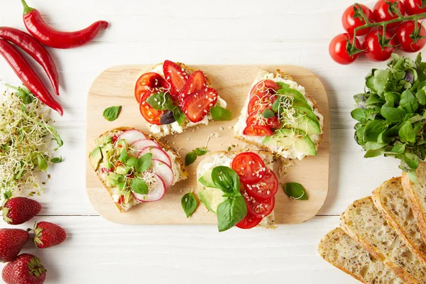 Vue du dessus des sandwichs végétariens sur planche à découper entourée d'ingrédients sur une surface en bois blanc — Photo de stock