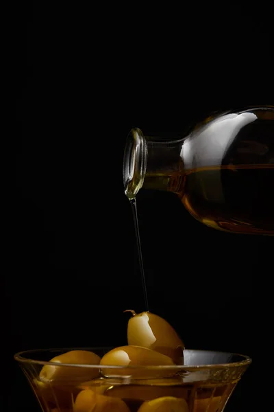 Ароматическое оливковое масло, льющееся в миску с оливками из бутылки, изолированной на черном — стоковое фото