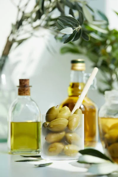 Bicchiere con cucchiaio e olive verdi, vasetto, varie bottiglie di olio aromatico con e rami su tavola bianca — Foto stock