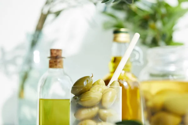 Primer plano de vidrio con cuchara y aceitunas verdes, frasco, varias botellas de aceite de oliva aromático con y ramas en bandeja de madera - foto de stock