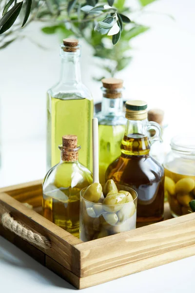 Verre avec cuillère et olives, bocal, diverses bouteilles d'huile d'olive aromatique avec et branches sur plateau en bois — Photo de stock