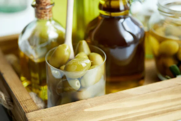 Primer plano vista de vidrio con cuchara y aceitunas verdes, frasco, varias botellas de aceite de oliva aromático en bandeja de madera - foto de stock