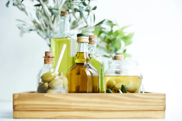 Vidrio con cuchara y aceitunas verdes, frasco, varias botellas de aceite de oliva aromático con y ramas en bandeja de madera - foto de stock