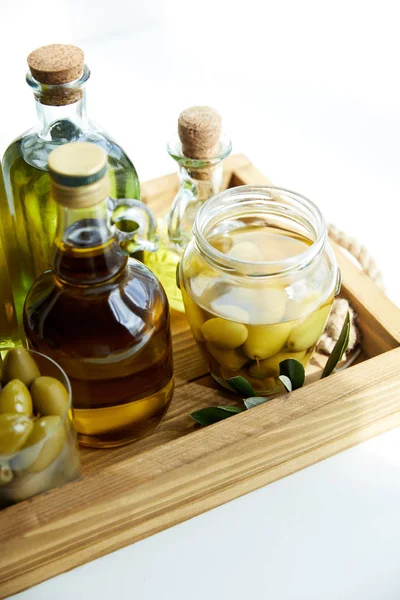 Frasco y vidrio con aceitunas verdes, varias botellas de aceite de oliva aromático con y rama en bandeja de madera - foto de stock