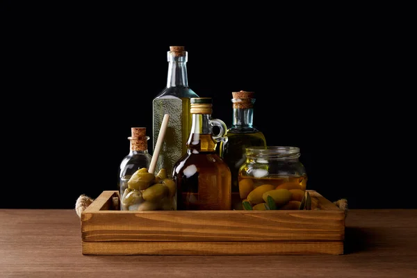 Various bottles of aromatic olive oil and jar with green olives on wooden table on black background — Stock Photo