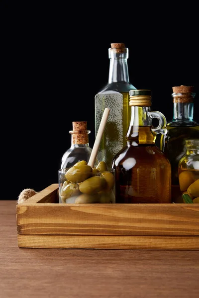 Different bottles of aromatic olive oil and jar with green olives on wooden table on black background — Stock Photo