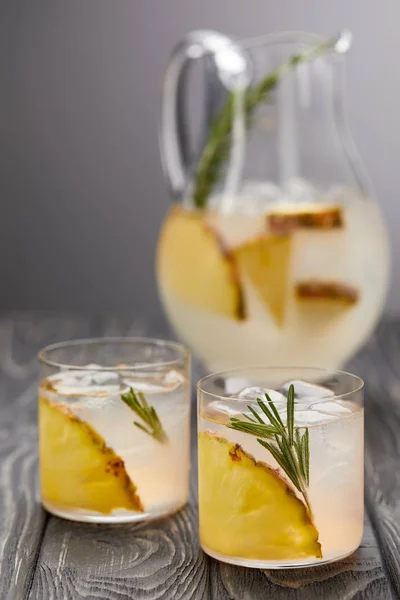 Jug of lemonade with pineapple and rosemary, two glasses of lemonade with pineapple pieces, ice cubes and rosemary on grey wooden tabletop — Stock Photo