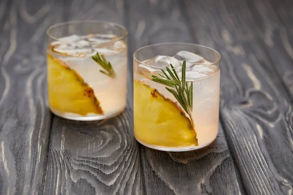 Two glasses of lemonade with pineapple pieces, ice cubes and rosemary on grey wooden tabletop — Stock Photo