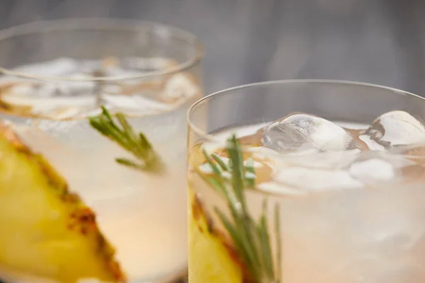 Enfoque selectivo de dos vasos de limonada con trozos de piña, cubitos de hielo y romero sobre mesa de madera gris - foto de stock