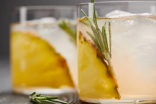 Selective focus of two glasses of lemonade with pineapple pieces, ice cubes and rosemary on grey wooden tabletop — Stock Photo
