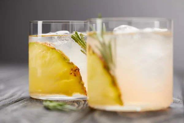 Два стакана лимонада с кусочками ананаса, кубиками льда и розмарином на сером деревянном столе — стоковое фото