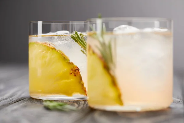 Two glasses of lemonade with pineapple pieces, ice cubes and rosemary on grey wooden tabletop — Stock Photo
