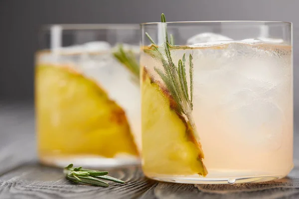 Selective focus of two glasses of lemonade with pineapple pieces, ice cubes and rosemary on grey wooden tabletop — Stock Photo