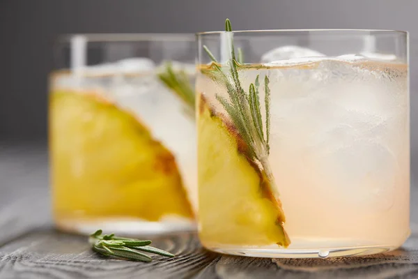 Foco seletivo de dois copos de limonada com pedaços de abacaxi, cubos de gelo e alecrim em mesa de madeira cinza — Fotografia de Stock