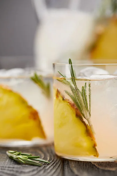 Jug with lemonade and two glasses of lemonade with pineapple pieces, ice cubes and rosemary on grey wooden tabletop — Stock Photo