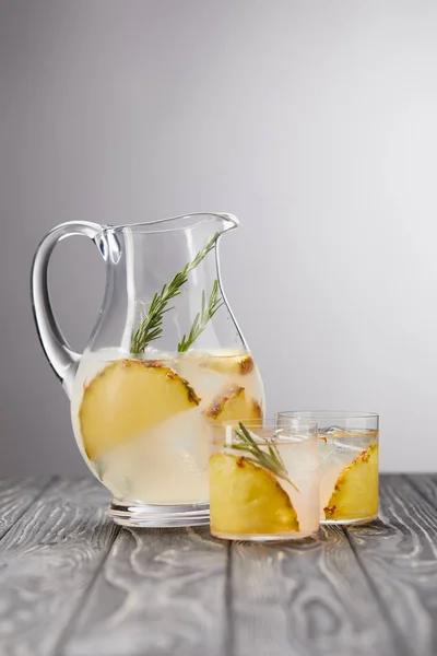 Jarro e dois copos de limonada com pedaços de abacaxi, cubos de gelo e alecrim em mesa de madeira cinza — Fotografia de Stock