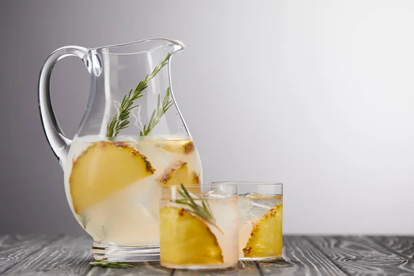 Krug und zwei Gläser Limonade mit Ananasstücken, Eiswürfeln und Rosmarin auf grauem Holztisch — Stockfoto