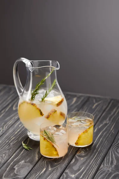 Krug und zwei Gläser Limonade mit Ananasstücken, Eiswürfeln und Rosmarin auf grauem Holztisch — Stockfoto