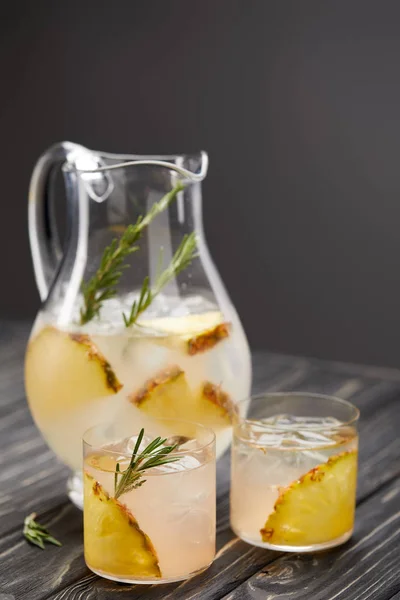 Krug und zwei Gläser Limonade mit Ananasstücken, Eiswürfeln und Rosmarin auf grauem Holztisch — Stockfoto