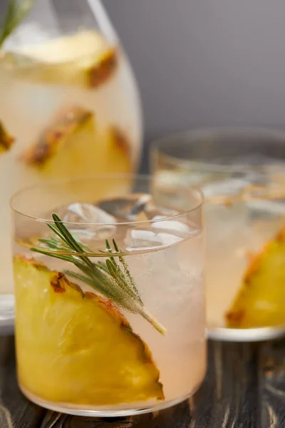 Selective focus of jug and two glasses of lemonade with pineapple pieces, ice cubes and rosemary on grey wooden tablet — Stock Photo