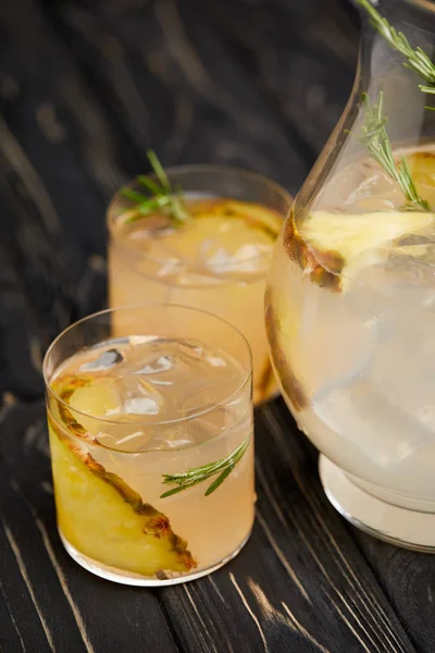 Vue partielle de la cruche et deux verres de limonade avec morceaux d'ananas, glaçons et romarin sur plateau en bois gris — Photo de stock